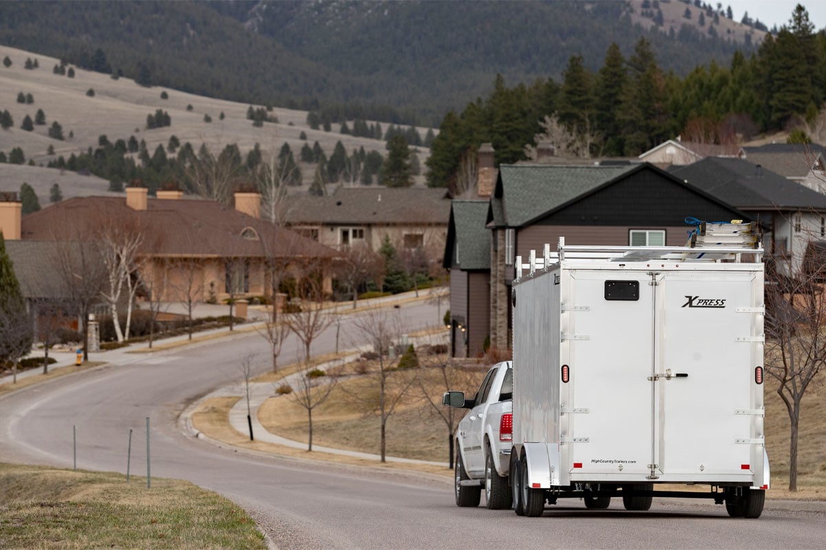 Back Side View Of White Truck Hauling White Xpress Enclosed Ultimate Contractor Trailer Through Residential Neighborhood